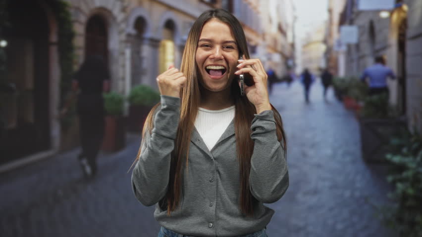 Woman brunette smiling holds smartphone to ear while pumping her fist on a narrow cobblestone street; joy celebration.