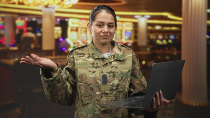 Woman soldier in camouflage holding laptop with palm up shrug gesture in casino building; uncertainty.