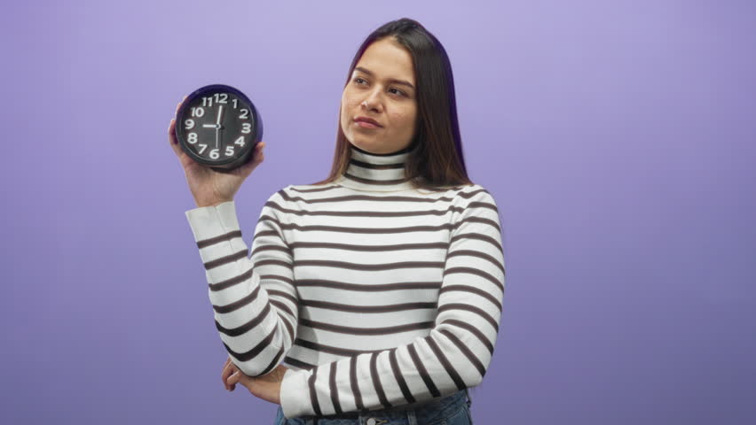 Woman brunette in striped turtleneck holding round clock with hand to chin against purple studio backdrop; thoughtful timing.