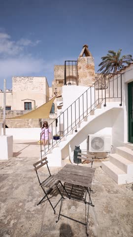 Static video of a woman walking up stairs to her terrace in southern Italy. Light wind gently moves the sun shade and palm tree, creating a calm Mediterranean slow living atmosphere and peaceful