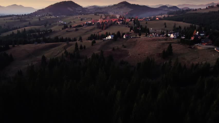 Aerial view of mountain village during sunset with glowing light over autumn hills and pine forest