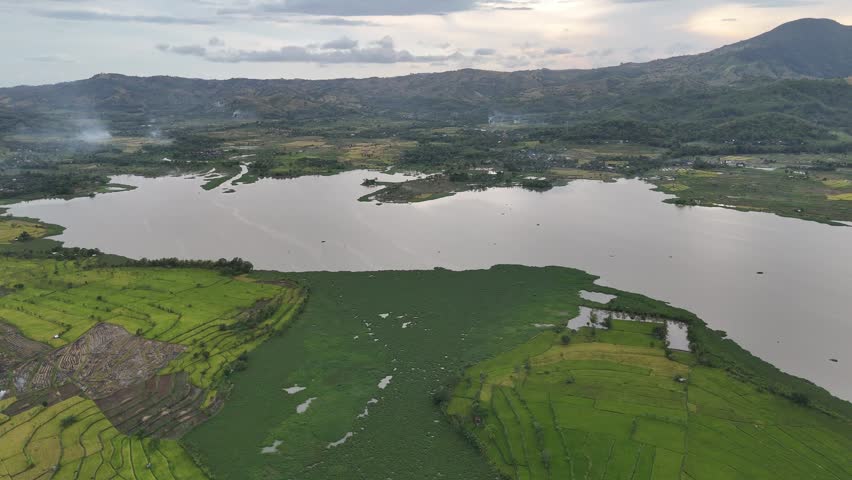 Aerial View of Lush Green Rice Terraces and Tranquil Lake Surrounded by Mountains under Overcast Sky