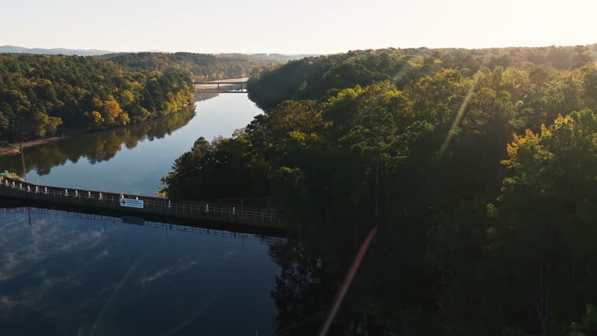 River channel below dam winding through dense forest under warm morning sunlight, aerial pan to reservoir with fog blowing across top of water