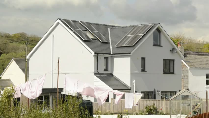 A modern, two-story white house with a dark grey roof featuring solar panels. The scene captures a bright day with a clothesline in the foreground holding laundry that is blowing gently in the wind. A wooden fence and a small greenhouse are visible in the backyard area, with a rolling green hill in the background under a soft, overcast sky.