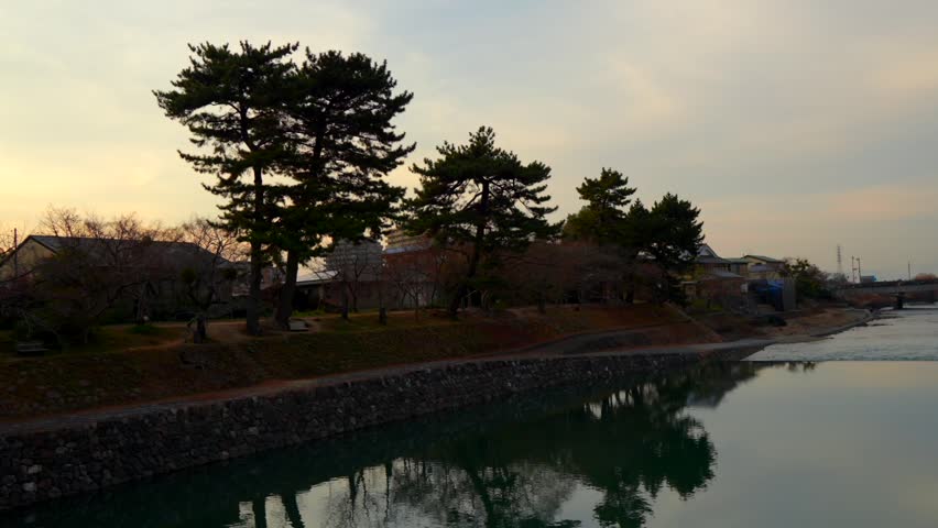 Static shot of a beautiful winter sunset over the Uji River, with its calm waters reflecting the sky and surrounding trees. Captured near the Asagiri Bridge in Uji, Japan.