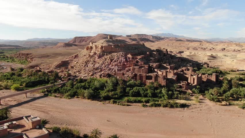 Aerial drone view of an ancient mud village built on a rocky hill surrounded by green oasis vegetation in the Atlas region of Morocco, showing traditional architecture within an arid desert landscape.