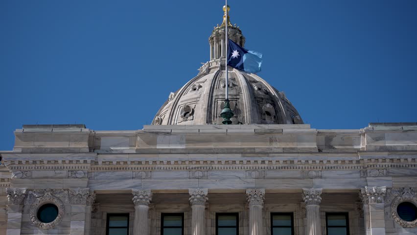 The flag of Minnesota flies on the dome of the State Capitol in Saint Paul under a bright blue sky.