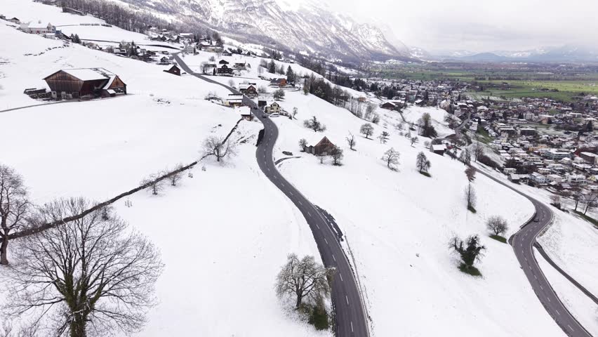 Drone flying over mountain road with snow on upper slopes and green valley below. Strong contrast between winter and spring in alpine landscape.