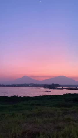 The footage captures a breathtaking sunset panorama over a calm lake in Central Java where the silhouettes of two massive volcanoes dominate the distant horizon. A thin silver crescent moon hangs delicately in the purple and orange twilight sky as the final glow of day reflects across the rippling water surface. Local wooden boats move slowly near the shore while the surrounding tropical landscape transitions into the quiet colors of dusk.

This scenic view showcases the natural majesty of the Indonesian volcanic arc during the golden hour in a peaceful rural setting. The composition features a foreground of wild grass and dark shoreline that leads the eye toward the vibrant gradient of the sky and the iconic peaks of Merapi and Merbabu. Soft natural lighting creates a serene atmosphere suitable for travel documentaries or cinematic background visuals.

A steady vertical shot emphasizes the height of the atmosphere and the vast scale of the mountains relative to the small fishing vessels on the reservoir. The clear sky allows for a sharp view of the celestial alignment above the volcanic range providing a sense of wonder and tranquility. This high-quality environmental footage represents the authentic beauty of Southeast Asian landscapes during a clear summer evening.