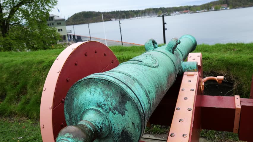 weathered green patinated historic cannon mounted wooden carriage points calm fjord waters forested shoreline sailboats nordic buildings form background norway toward coast