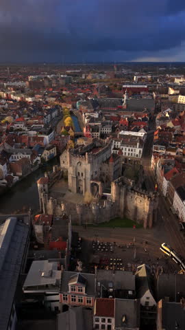 Gravensteen castle aerial golden hour, dramatic storm clouds, warm sunlight grazing stone battlements, empty market square, cobbled streets, surrounding red tile rooftops, cinematic drone
