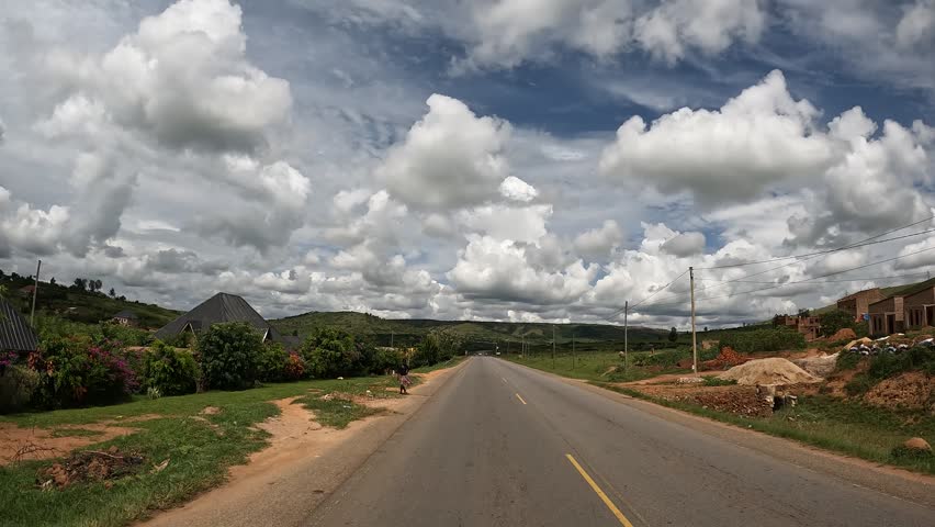 POV footage driving along a paved highway through the rolling green hills and rural countryside of Republic of Uganda under a cloudy sky.