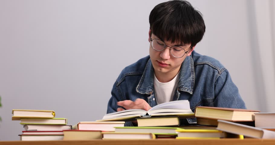 Student in glasses with many books preparing for exam at desk indoors