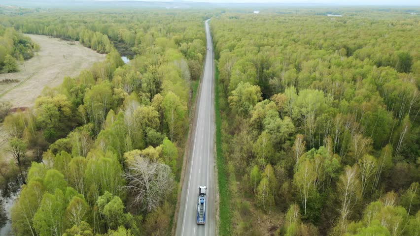 Aerial view shows a long straight road. Green trees line both sides of the road. Single truck travels down the road. No people or animals are visible. Perfect for nature, travel