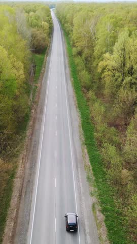 Aerial view of a solitary car on a long, straight road. Trees line both sides, green and full of spring. The car moves slowly, centered on the lane. Road stretches into distant horizon, peaceful