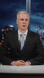 Experienced Male Anchor in Classical Suit Presenting News, Looking at Camera And Talking at Desk With Documents in Studio With Breaking News Display. Corporate Media Presentation. Vertical Shot. - Powered by Shutterstock - Get 15% off with code: PIKWIZARD15