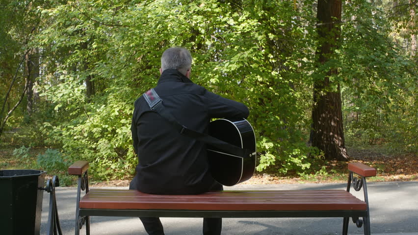 Older white man sitting bench playing guitar gray hair, black jacket, urban park with tall trees and dappled sunlight, solo street musician practice, reflective mood, wooden bench, paved