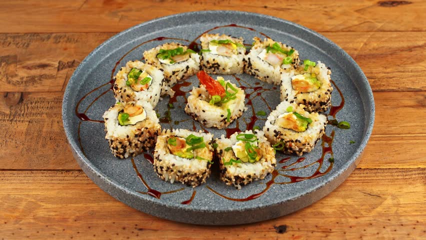 A plate of sushi rolls with sesame seeds and sauces is lifted from a wooden table, showcasing gourmet food presentation