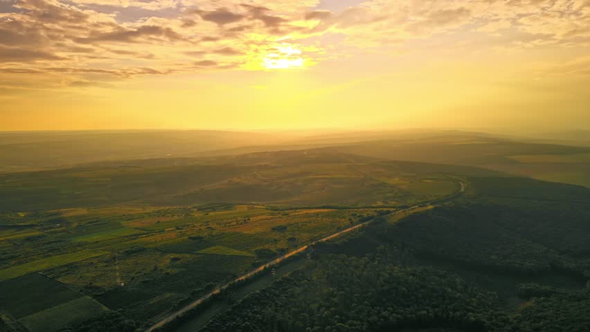 Golden sunrise illuminates vast landscape with rolling hills and forest, aerial drone view footage.