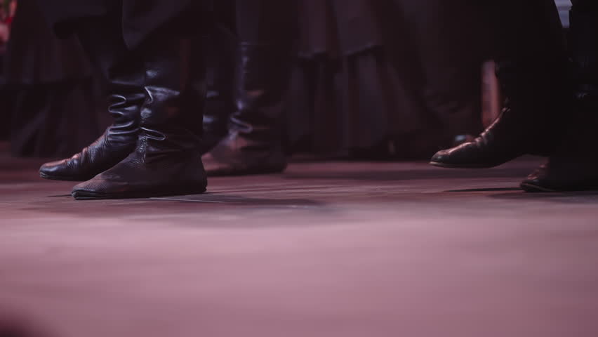 Low-angle close-up of synchronized dancers black leather boots striking a stage under dramatic red lighting, capturing rhythmic footwork and ensemble choreography in a dynamic theatrical folk