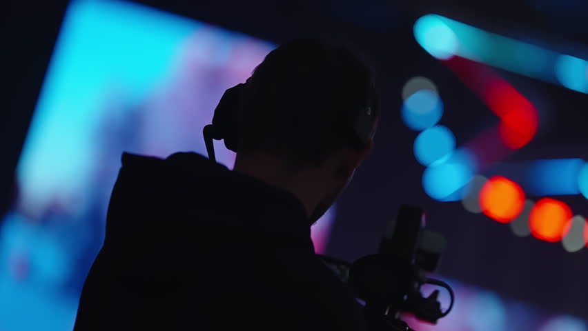 Silhouette of a camera operator filming on a vibrant stage with colorful bokeh lights and neon shapes, capturing live event action in a dark venue with dramatic backlighting.
