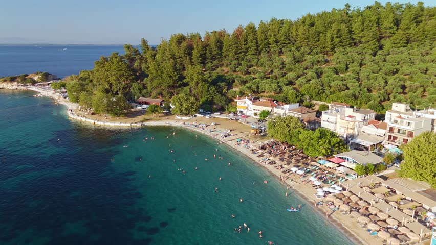 Aerial view of a crowded Limenas beach with crystal clear turquoise water of Aegean sea, green trees, and sunbeds under a blue summer sky on Thasos island, Greece