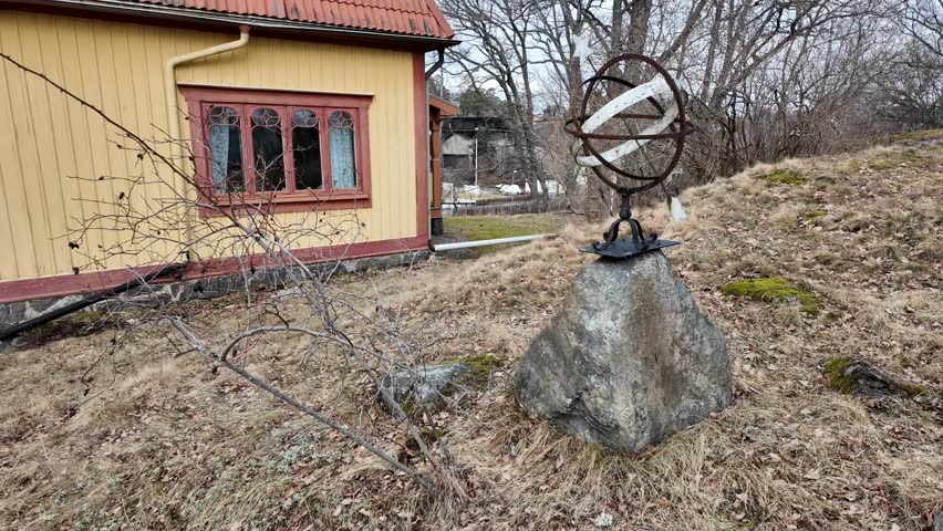 A historic outdoor display in a public open-air museum in Sweden, captured in early spring 2026. The scene features a traditional yellow wooden building with decorative windows and a tiled roof, typical for Swedish countryside architecture. In the foreground stands a metal astronomical armillary sphere mounted on a stone base, adding a scientific and historical element to the setting.

The ground is covered with dry grass, fallen leaves, and patches of moss, while leafless trees surround the area, indicating the early spring season. Soft daylight and a partly cloudy sky create natural lighting and highlight textures of wood, metal, and stone.