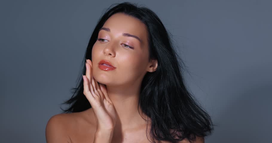 Close-up shot of attractive young woman with flawless skin gently touching her face in gray studio setting. Soft lighting highlights natural beauty. Ideal for skincare, cosmetics, and beauty product