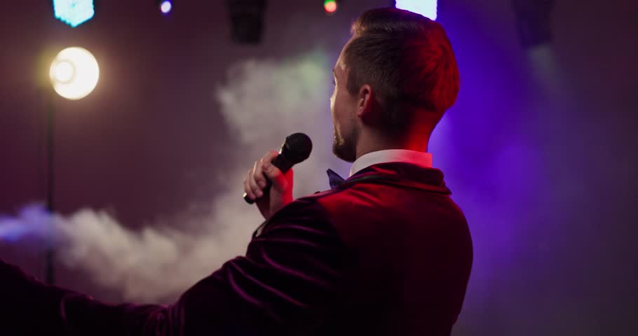 Host presenter showman with microphone on stage. Emcee and speaker engages crowd under lights and haze, ideal for concert promos, events, and nightlife ads. Clear show hosting and presentation.