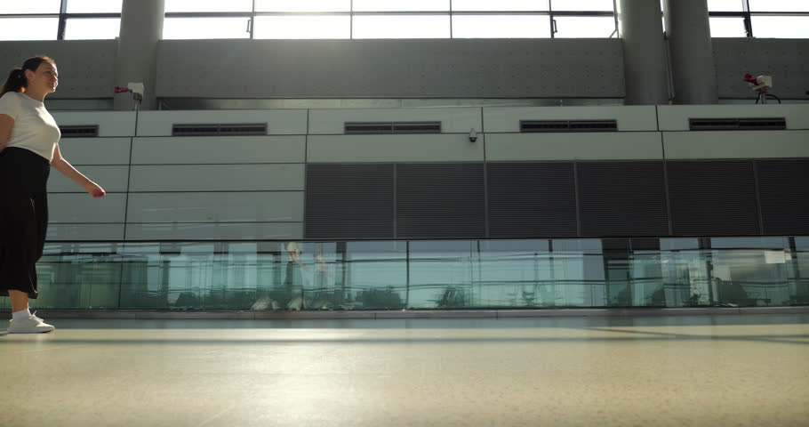 Side slow motion view from low angle of female passenger walking through empty sunlit airport terminal, pulling her carry on luggage on her way to the departure gate for her next business trip or vacation