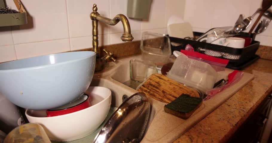 Male hands washing a wooden item in a messy kitchen sink full of dirty dishes