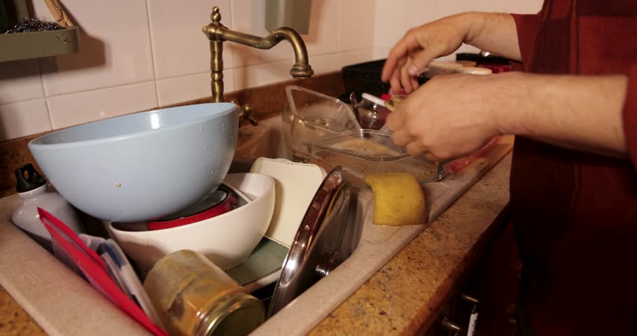 Unrecognizable man doing daily household chores by washing dirty dishes in a sink full of plates