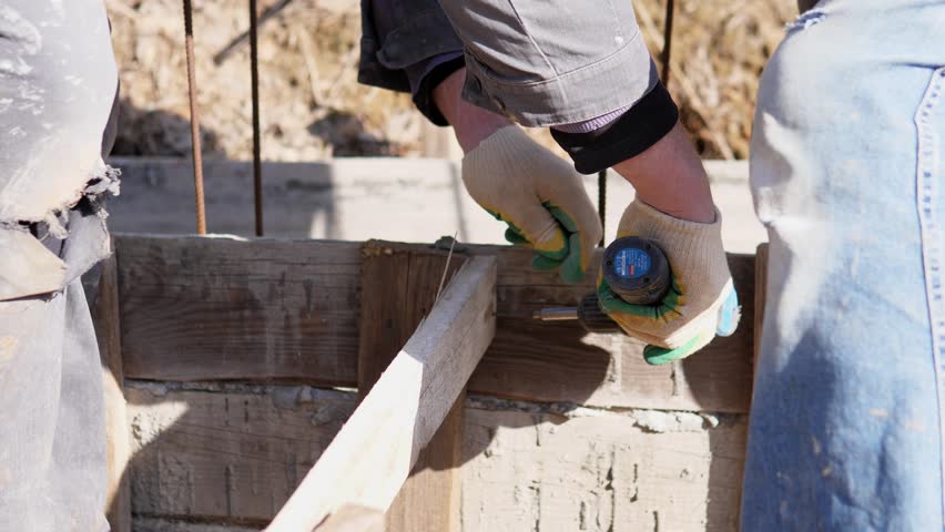 Closeup Gloved Hands Fastening Plank To Concrete Block With Cordless Drill, Screw Insertion And Firm Grip, Masonry Form Detail, Skilled Builder Focusing On Alignment, Dusty Outdoor Site,