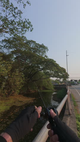 First person perspective of an angler casting a fishing rod over a small canal from a roadside bridge during warm golden hour light. Hands holding the rod and reel in action, capturing outdoor fishing lifestyle, adventure, and peaceful countryside environment in Southeast Asia.