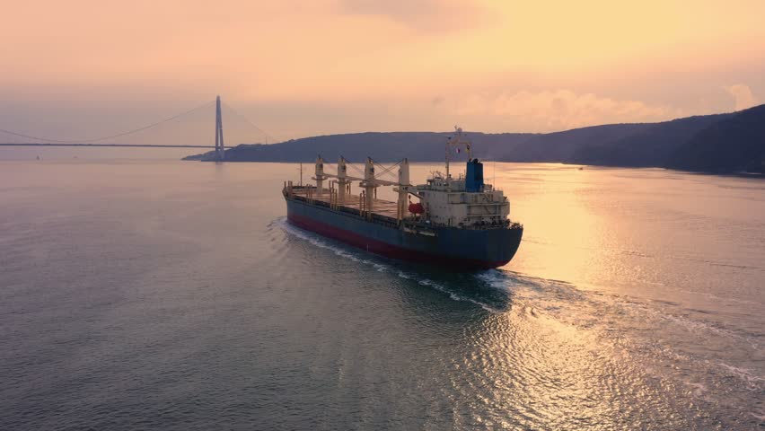 Bulk carrier deck and cargo cranes filmed in close pursuit at golden hour, warm sunset sky glowing over calm waters with suspension bridge silhouette in the distance