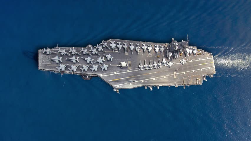 View the aircraft carrier with a full deck of fighter jets in a vibrant blue ocean, representing military power and readiness in ongoing geopolitical tensions.