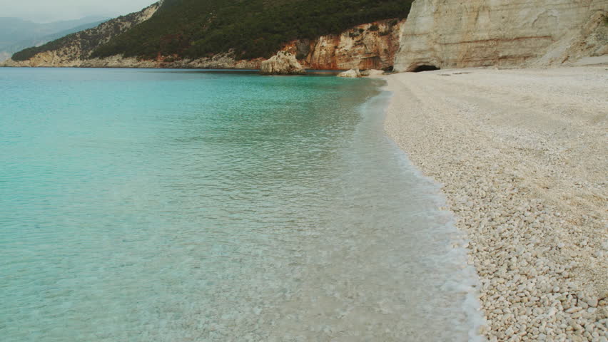 Crystal clear turquoise waves lapping pebble shoreline below green cliffs in Kefalonia Greece