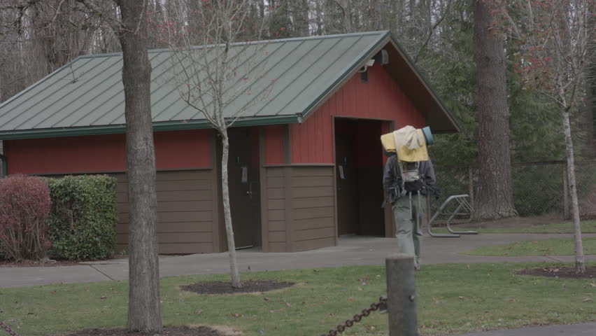 Hiker with a backpack and camping gear walking towards a public restroom in a wooded park area