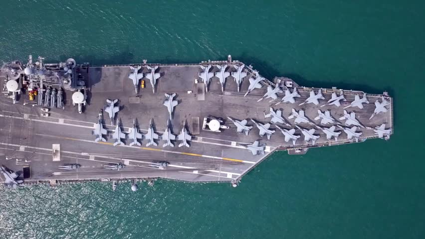 High-angle aerial shot of a military aircraft carrier featuring multiple fighter jets and naval personnel in formation on the deck. Ideal for defense, maritime, aviation, and military-themed projects.