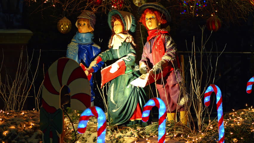 Three vintage Christmas caroler statues with glowing candy canes. Outdoor holiday display of singing figures in winter attire with snow at night in New York City.