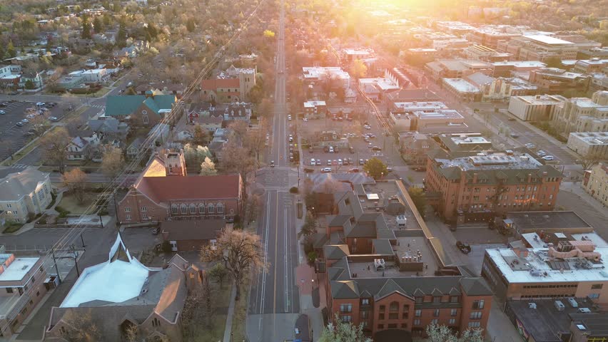 Broadway corridor in central Boulder stretches past churches, small shops, branching residential streets below warm sunrise light. Shingles, flat decks, parked cars, leafless canopies by roadway. USA