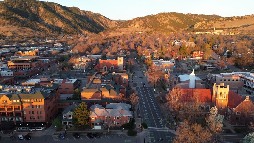 Downtown Boulder, Pearl Street corridor sunlit foothills, early spring trees Front Range. Warm golden light skims brick roofs, pale stucco walls, bare branches, patchy forested slopes ridgelines. USA