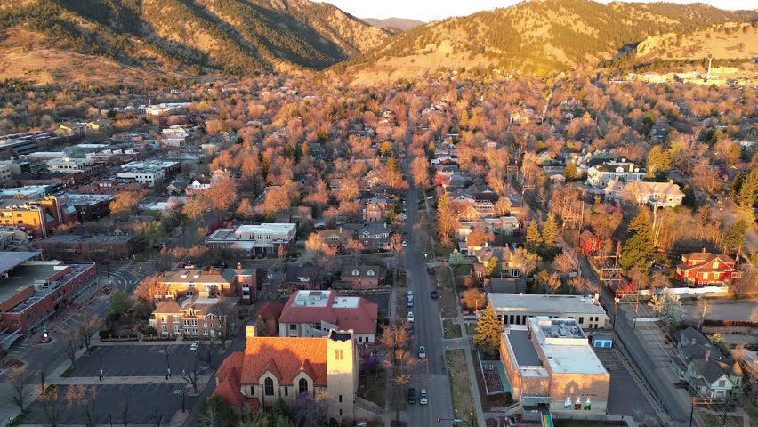 Central Boulder Street grid, church tower, low commercial blocks extend toward rising Front Range foothills. Brick facades, flat roofs, leafless treetops, rocky slopes layer distant ridgelines. USA
