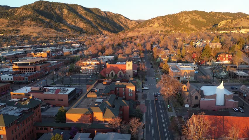 Historic downtown Boulder streets frame red roofed church tower, white roofed hall, compact blocks, Front Range slopes. Warm light glows over shingles, bare trees, mixed masonry by foothill ridges. US