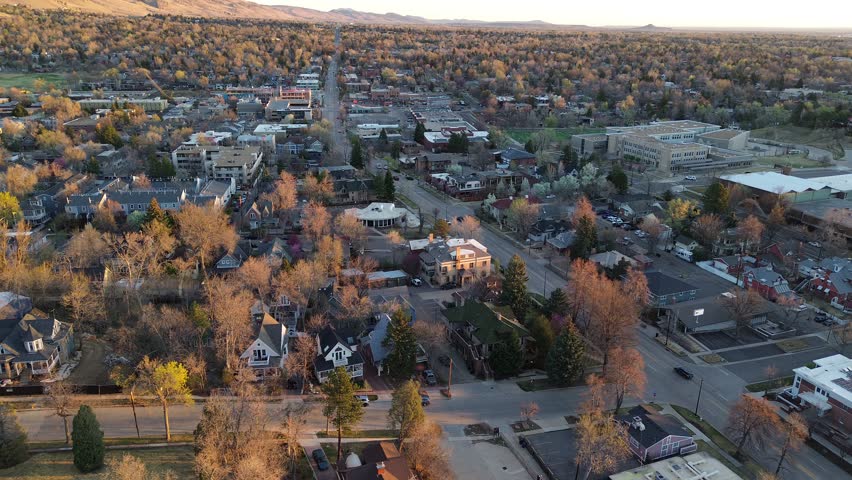 Mixed Boulder residential, small commercial blocks form loose grid beneath distant foothill ridges, tree lined streets. Warm light washes gabled roofs, flat decks, parked cars, bare canopies. USA