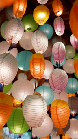 Many colorful glowing paper lanterns hanging from ceiling indoors. Electrical round and fish-shaped handmade paper lanterns garlands used as popular decoration for streets and indoor areas in Vietnam.