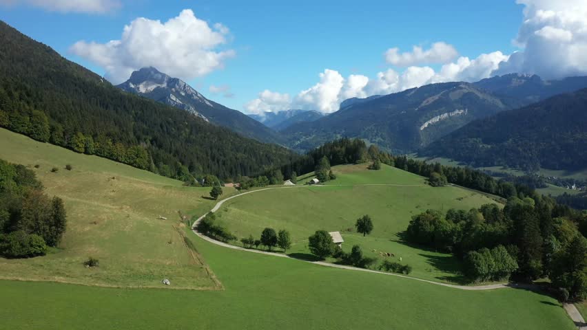 Aerial view of rolling green hills, forests, and mountain peaks in the Massif de la Chartreuse, France, under a blue sky with clouds.