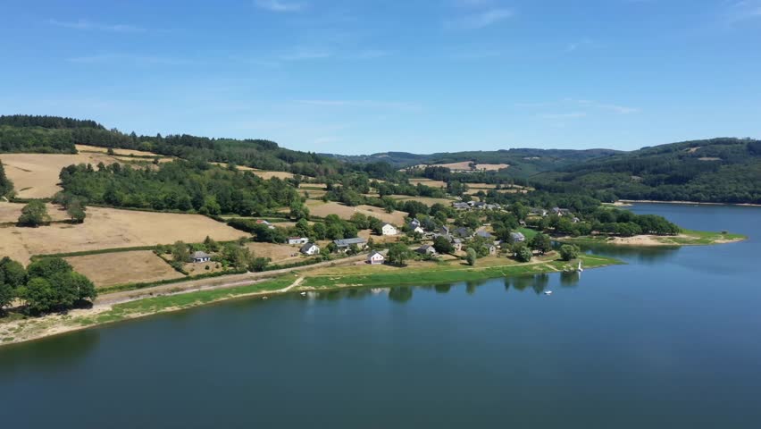 Aerial view of the Panneciere Chaumard Reservoir with a village, green hills, and calm water under a clear blue sky in Burgundy, France.