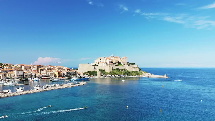 Scenic view of Calvi citadel and marina with yachts and boats on the clear blue Mediterranean Sea in Corsica, France under a bright sky.