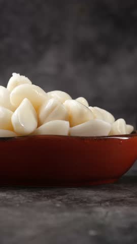 Close up slow motion of peeled garlic cloves piled in a rustic red bowl on dark stone surface.